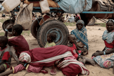 A family of Sudanese refugees who have been displaced by the ongoing conflict. Four children sitting on the ground along with two women, while one is lying on the ground. On the background is a horse cart with their belongings.